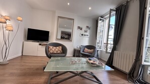 A modern living room with a glass coffee table, two armchairs, a flat-screen TV, a large mirror, and a window with curtains. - Charming Apartment in Montmartre (Paris)