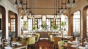Un restaurant doté de nombreuses tables, de chaises, de luminaires suspendus et de grandes fenêtres ornées de plantes.