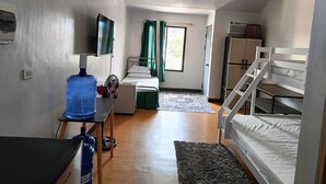 A compact room with a bunk bed, a desk with a water dispenser, a small table, a fan, and a window with curtains.