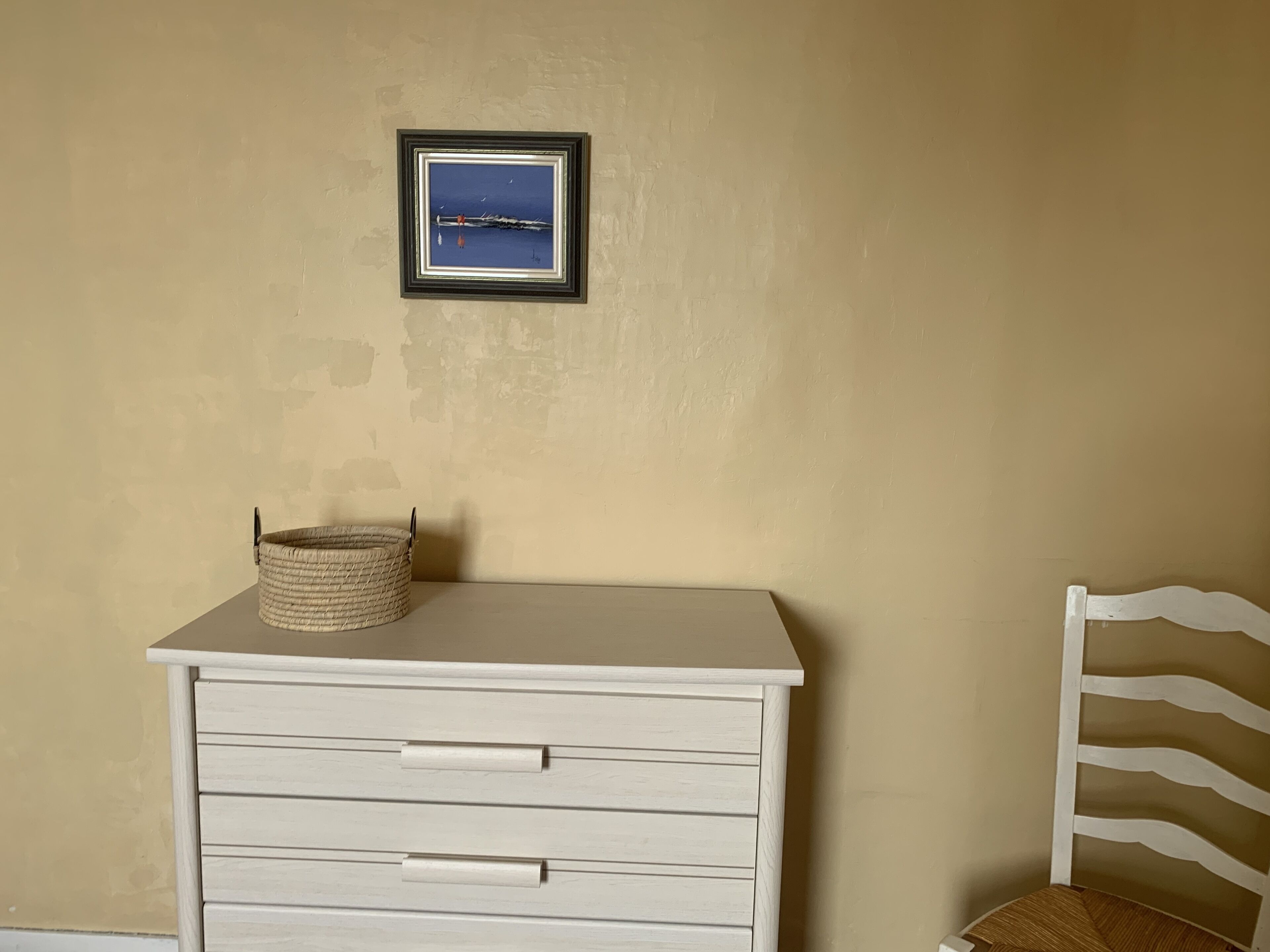 A white dresser with drawers, a framed picture on the wall, and a wicker basket on top.
