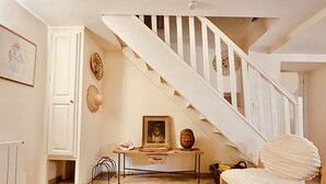 A staircase with white railings leading to an upper floor, a chair with a white cushion, a framed picture on a small table, and a tiled floor. - Charming Gîte in the Heart of Beaujolais (Lancié)