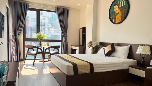 A hotel room with a large bed, a desk with a chair, a window with curtains, and a painting on the wall. - Au79 Hotel & Apartment (Hai Phong)