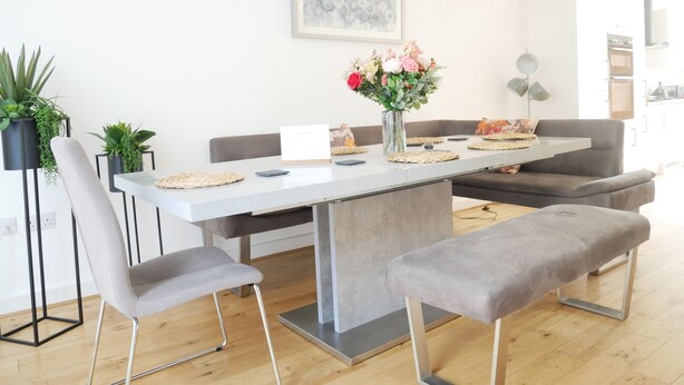 A modern dining area with a white table, grey chairs, and a bench. There are potted plants and a vase with flowers on the table. - Town House (Ulverston)