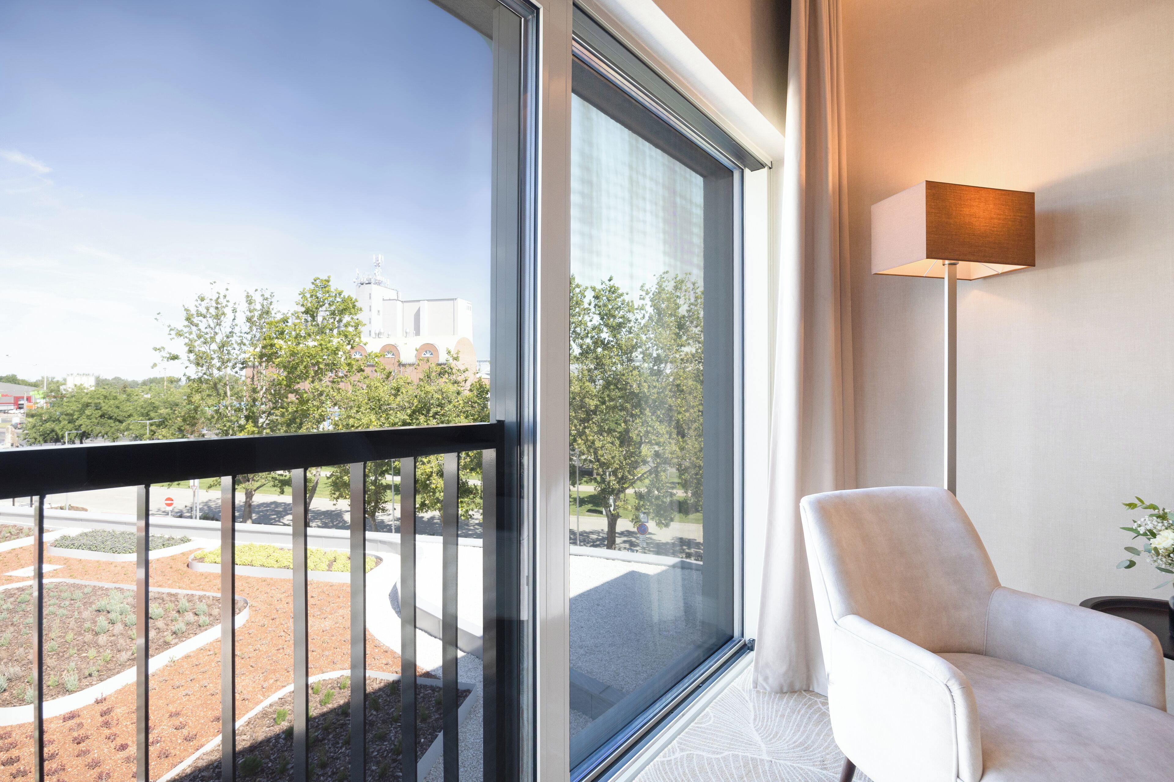 A hotel room with a large window offering a view of a landscaped area and a building in the distance. There is a white armchair and a floor lamp.