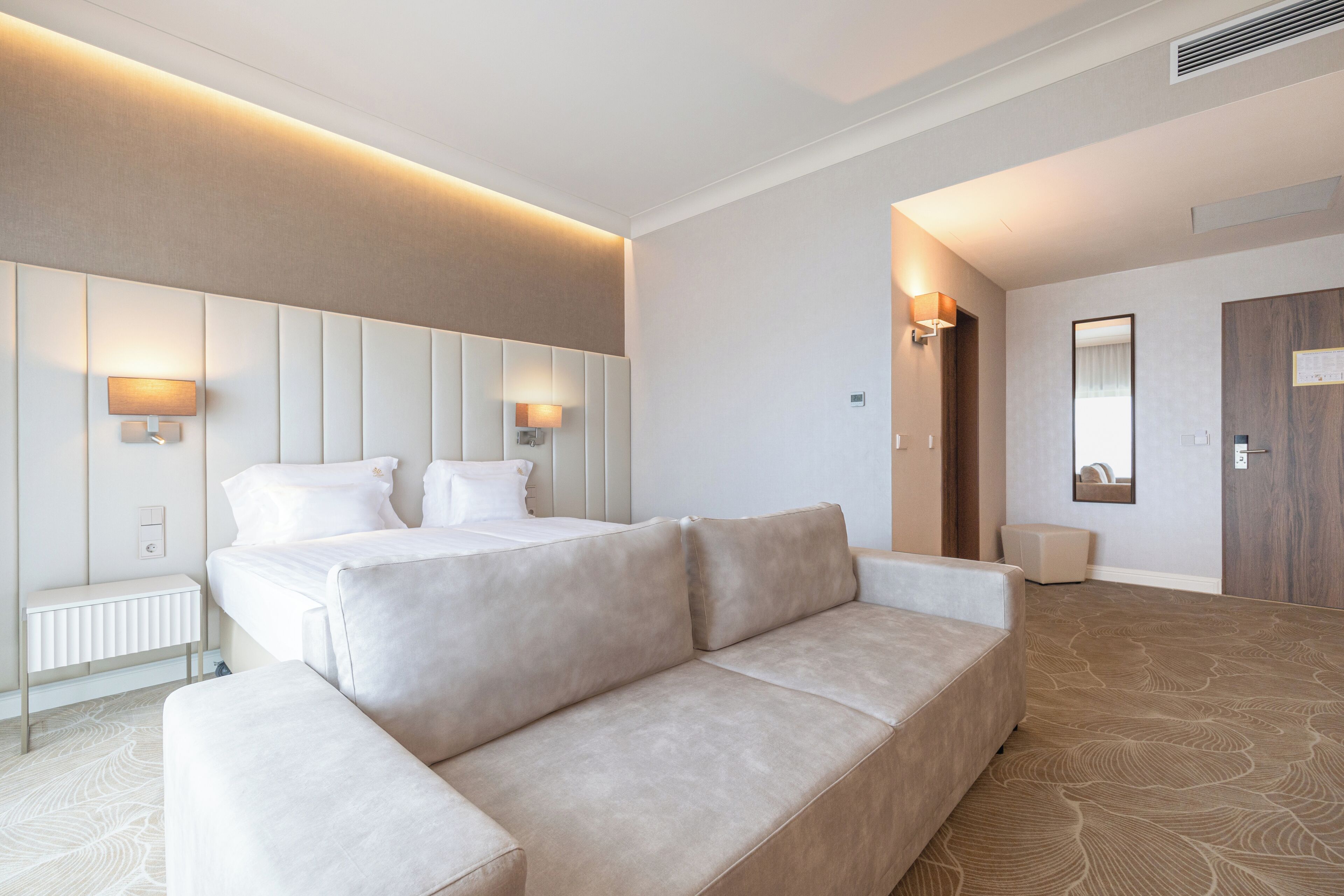 A modern hotel room with a large bed, two bedside lamps, a sofa, and a mirror.