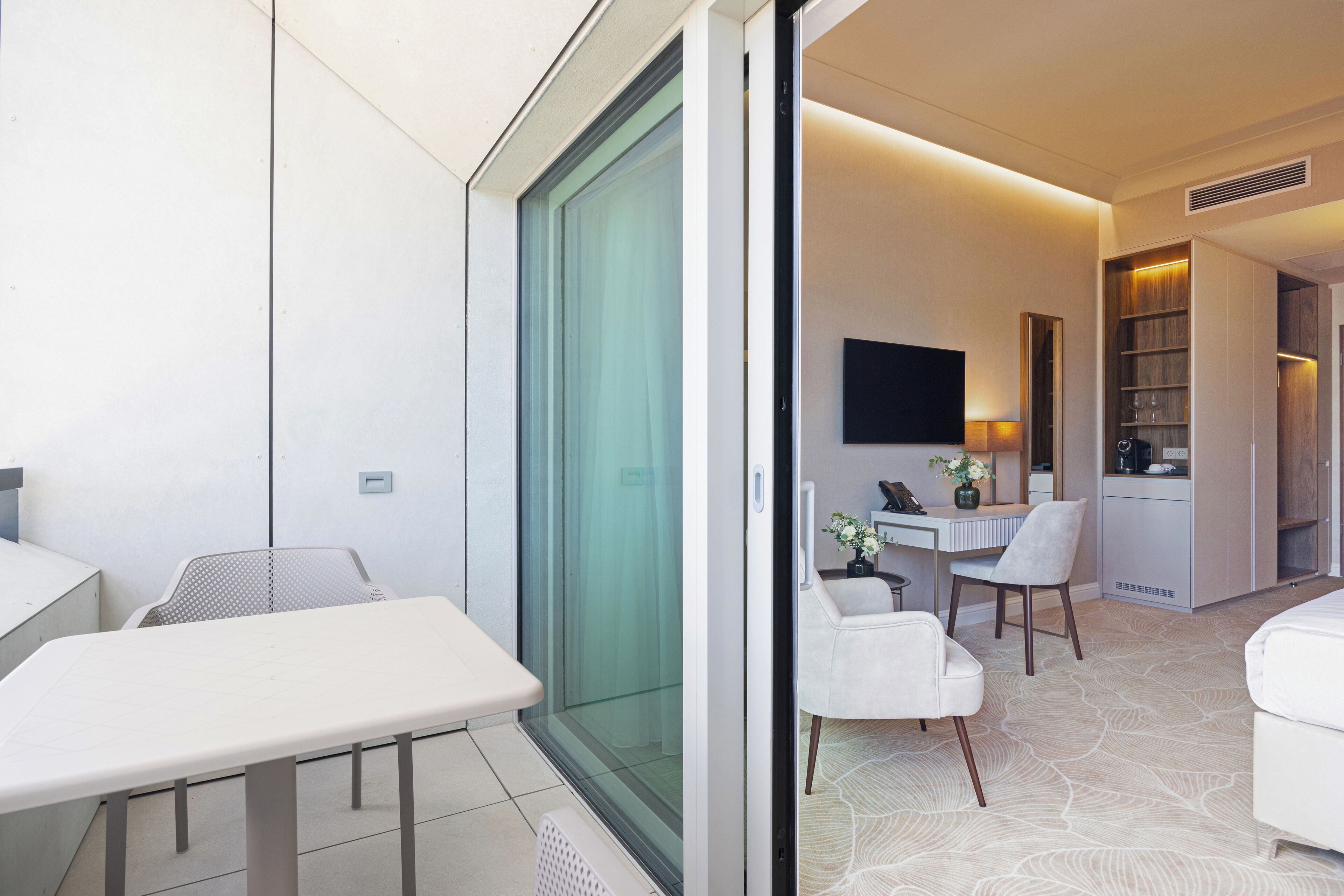 Una habitación de hotel moderna con puerta corredera de vidrio, un área de comedor con mesa y sillas, un televisor de pantalla plana montado en la pared y una estantería empotrada.