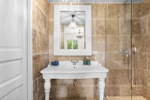 A bathroom with a white pedestal sink, a mirror with a decorative frame, a chandelier, and a shower with a glass door. - Caribbean Lofts 61 - Waterfront (Kralendijk)