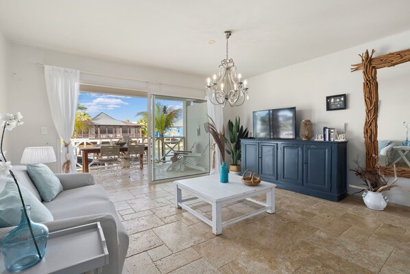 A spacious living area with a sofa, coffee table, and a TV on a cabinet. There is a balcony with a dining set and a view of the outside. - Caribbean Lofts 61 - Waterfront (Kralendijk)