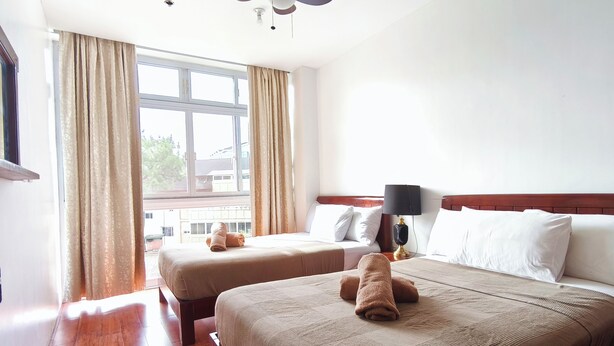 A hotel room with two beds, a ceiling fan, a window with curtains, and a view of buildings outside. - Grandview Residences by Vilma near Burnham Park-204 (Baguio)