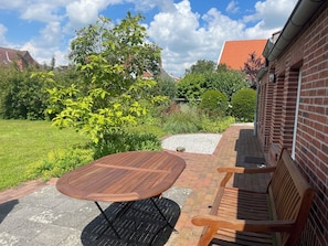 A patio with a wooden table and benches, surrounded by a brick wall and greenery. - Vacation home with garden (Krummhörn)