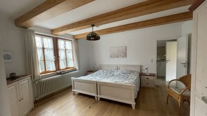 Un dormitorio con piso de madera, una cama con colcha estampada, una ventana con cortinas, un radiador, techo de madera y una puerta que da a otra habitación.