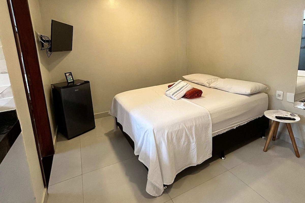 A modern hotel room with a single bed, a small bedside table, a flat-screen TV mounted on the wall, and a portable heater on the floor.