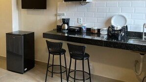 A compact kitchen with a microwave, coffee maker, and a sink. There are two black bar stools and a black refrigerator. - Morada do Sol Suítes (Arraial do Cabo)