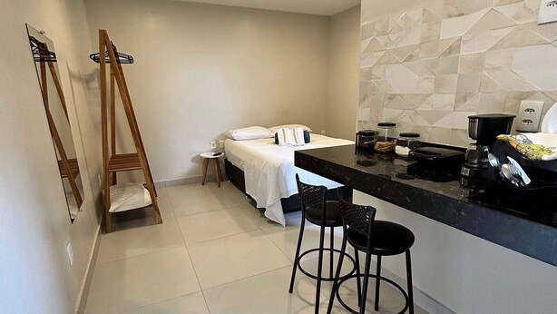 A modern hotel room with a bed, a small sitting area with two chairs, a kitchen counter, and a large mirror. - Morada do Sol Suítes (Arraial do Cabo)