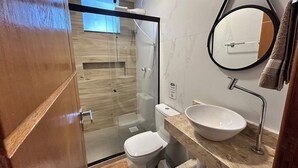 A modern bathroom with a glass-enclosed shower, a toilet, a sink with a marble countertop, and a mirror with a towel hanging on the wall. - Morada do Sol Suítes (Arraial do Cabo)