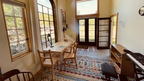 A cozy room with a wooden table, chairs, a patterned rug, a piano, and large windows offering views of the outdoors.