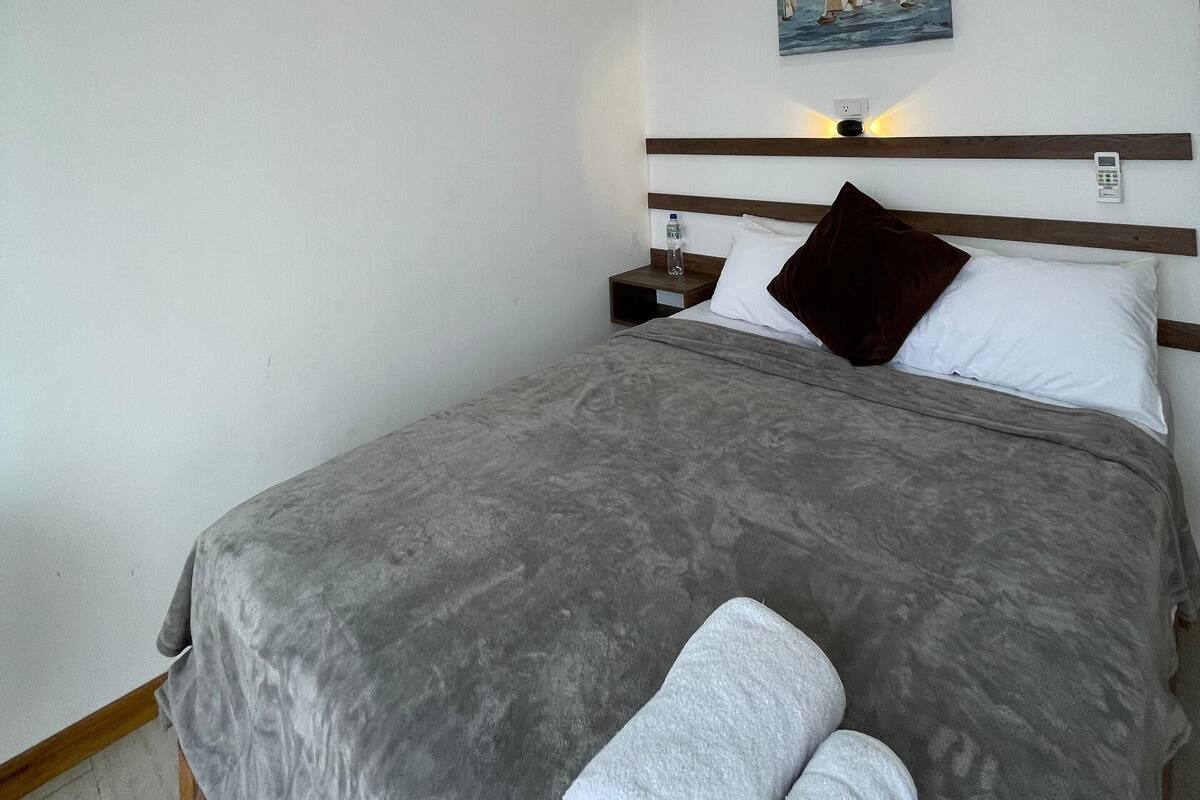 Een net opgemaakt bed met een grijze deken en twee witte handdoeken, een schilderij van zeilboten aan de muur, en een nachtkastje met een waterfles.