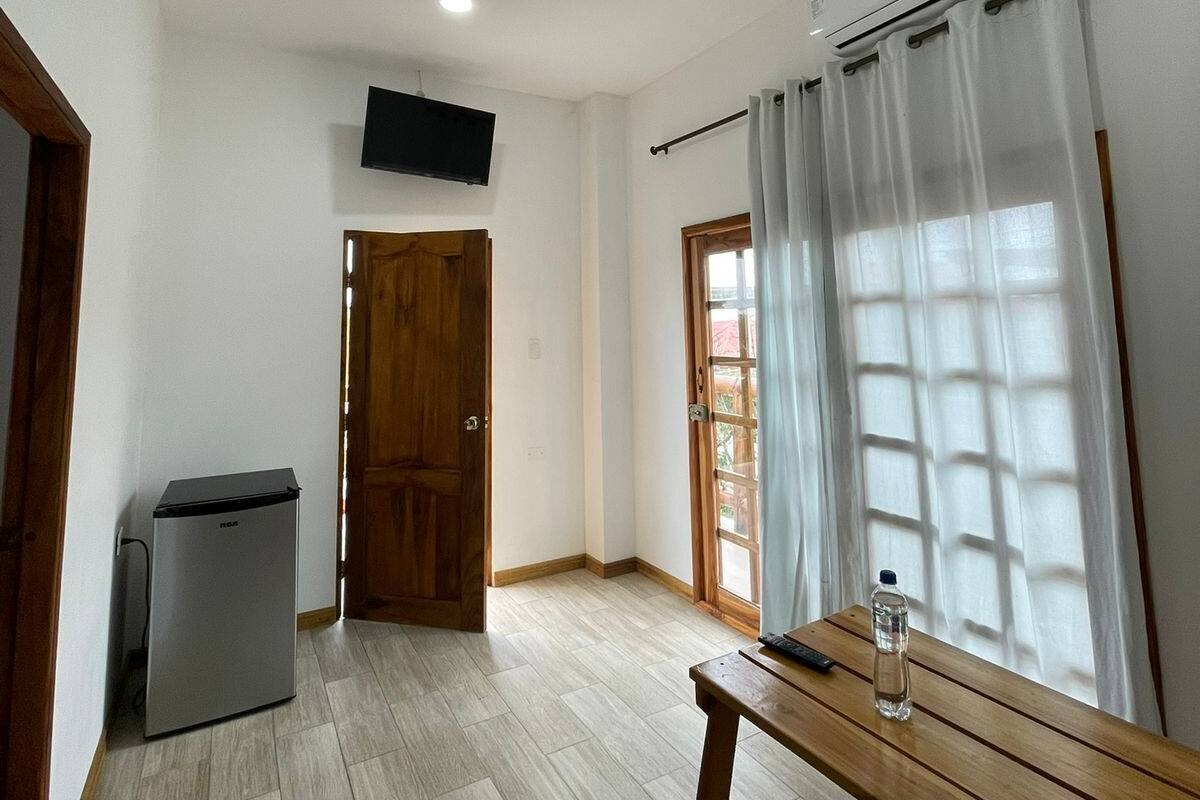 Een kamer met een houten bankje, een tafeltje met een fles, een tv aan de muur en een plafondventilator.