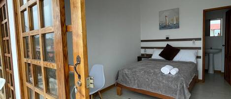 Un dormitorio con una cama, una silla, un cuadro y un baño visible a través de una puerta abierta.