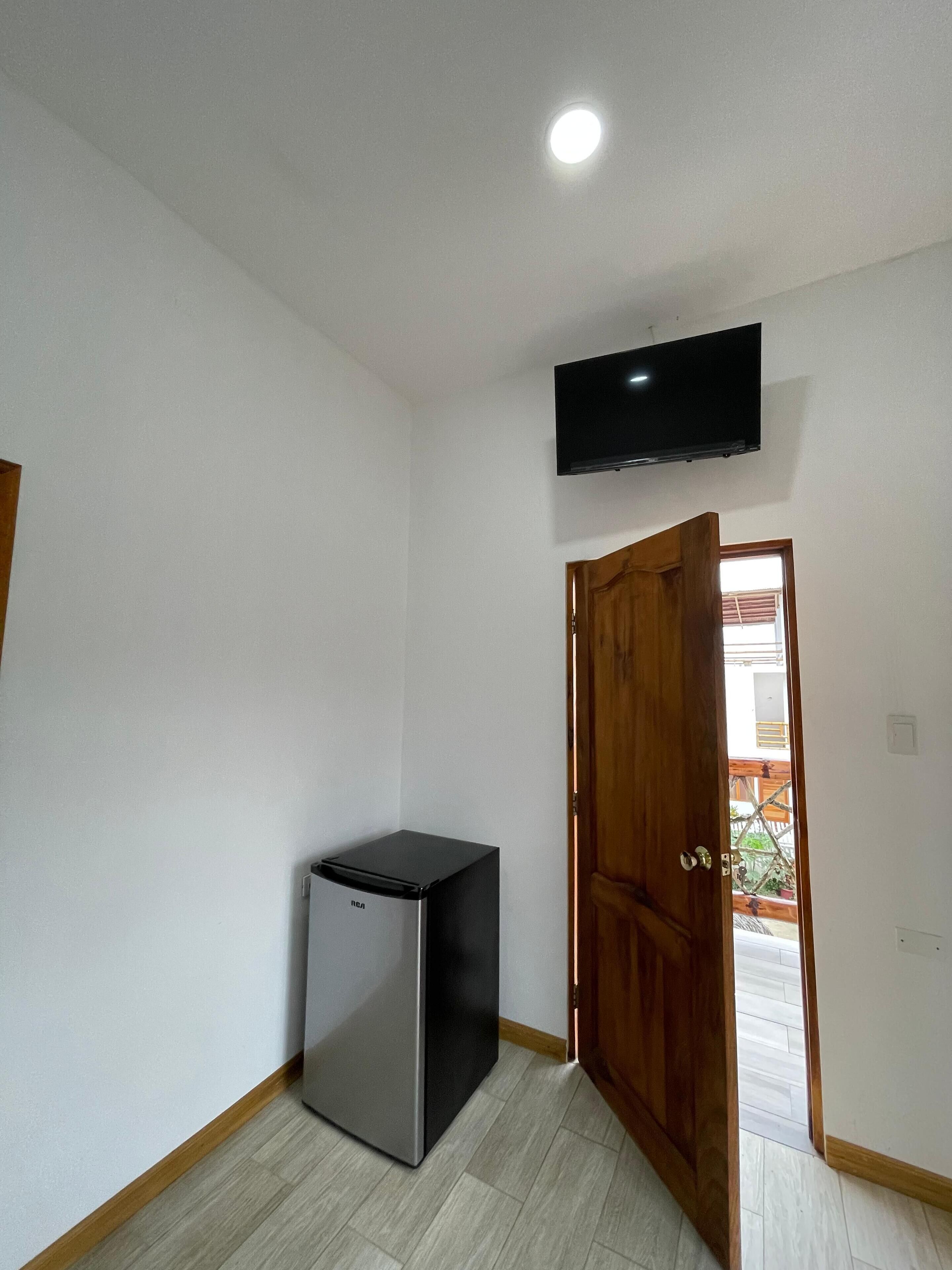 Una habitación pequeña con refrigerador, televisor montado en la pared y puerta de madera que da a un espacio exterior.