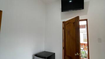 Una habitación pequeña con refrigerador, televisor montado en la pared y puerta de madera que da a un espacio exterior.