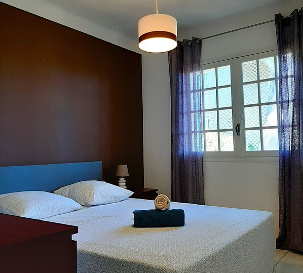 A bedroom with a large bed, a bedside table with a lamp, a window with curtains, and a hanging light fixture.
