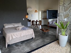 Quarto moderno com uma cama grande, área de refeições com cadeiras, uma mesinha e um vaso de planta.