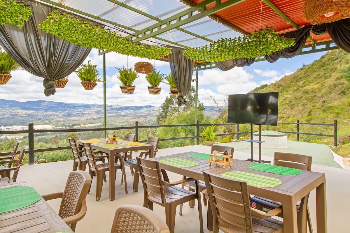 Un patio exterior con mesas y sillas, rodeado de plantas colgantes y vistas al campo.