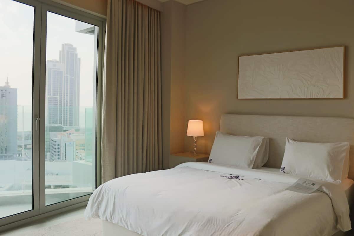 Ein Hotelzimmer mit einem großen Bett, einem Nachttisch mit Lampe, Blick auf die Stadt durch ein großes Fenster und einem Bild an der Wand.