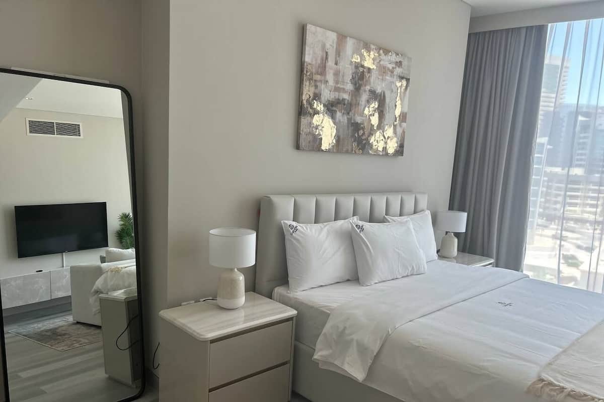 Ein modernes Hotelzimmer mit einem großen Bett, einem Flachbildfernseher, einem Nachttisch mit Lampe und Blick auf die Stadt durch das Fenster.