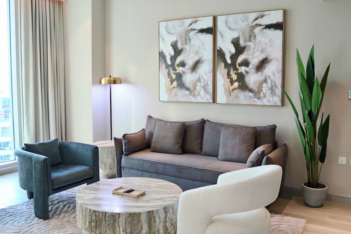 Ein modernes Wohnzimmer mit einem grauen Sofa, einem weißen Sessel und einem runden Couchtisch. An den Wänden hängen zwei abstrakte Gemälde und in einem Topf steht eine große, grüne Pflanze.