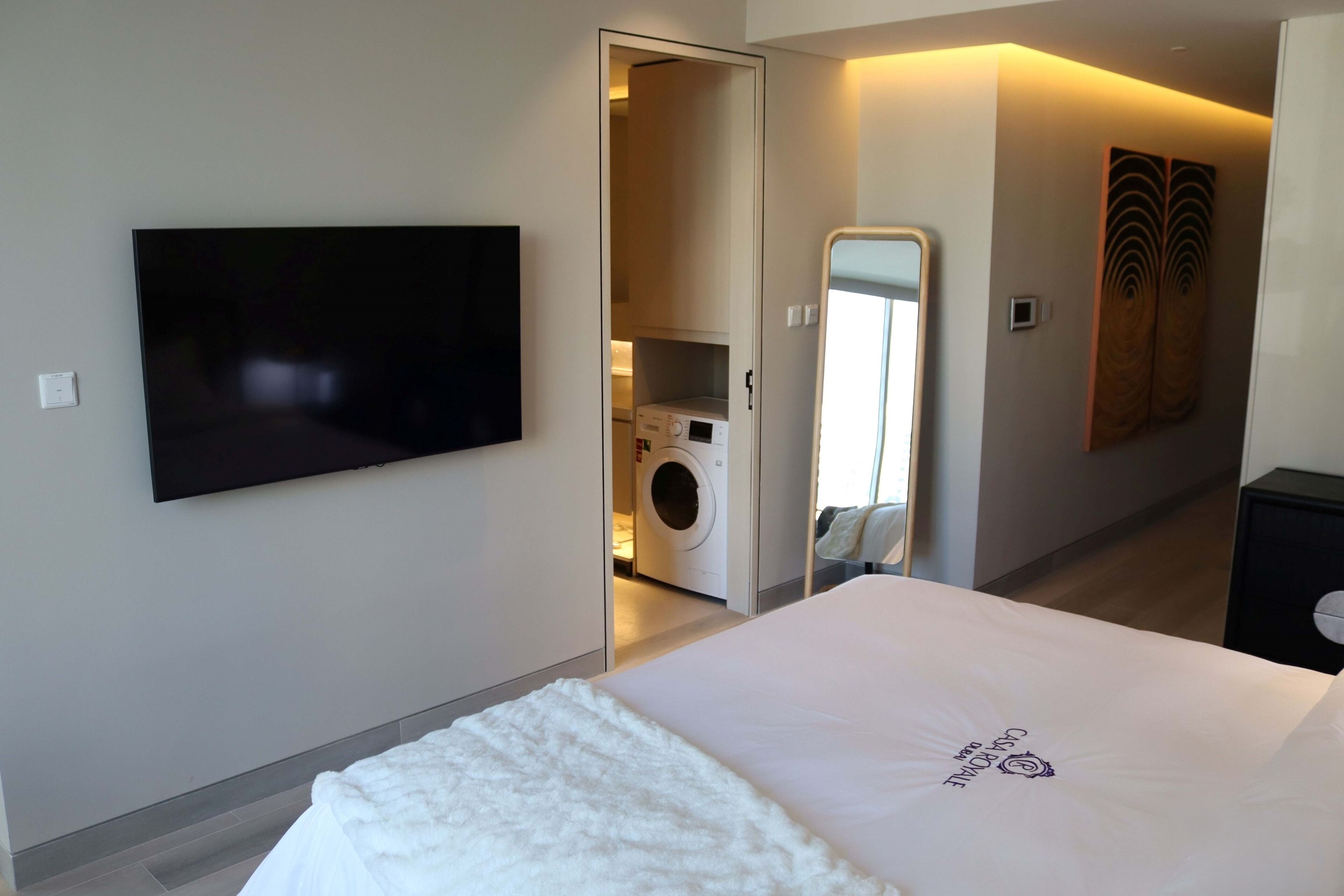 A hotel room with a bed, a TV mounted on the wall, a washing machine, and a mirror.