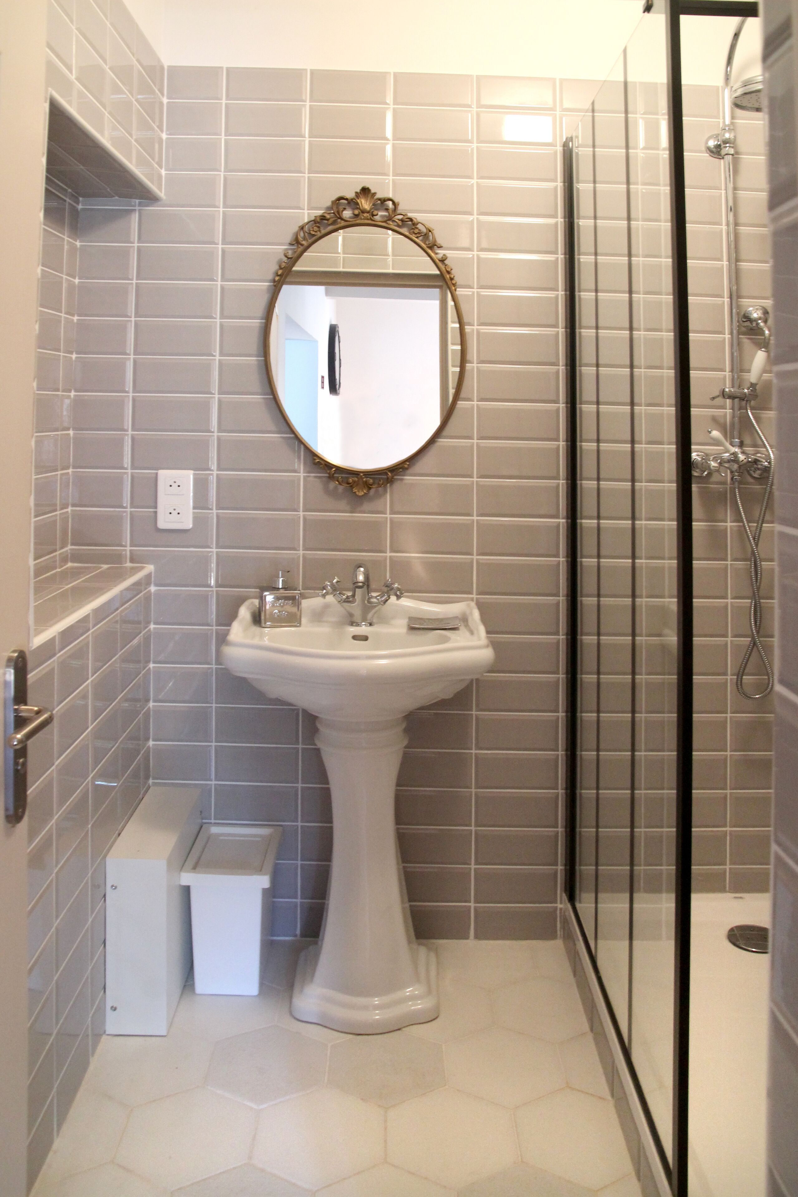 A bathroom with a pedestal sink, a mirror with an ornate frame, a shower enclosure, and a small trash bin.