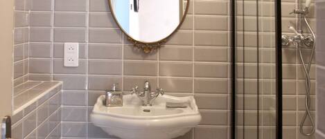 A bathroom with a pedestal sink, a mirror with an ornate frame, a shower enclosure, and a small trash bin.
