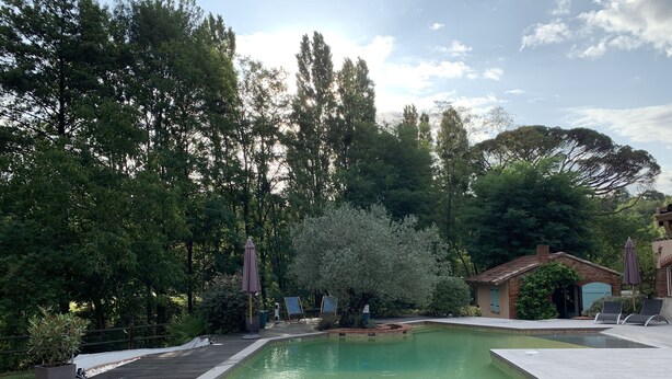 Una piscina con un deck circundante, rodeada de árboles y un pequeño edificio de ladrillo.