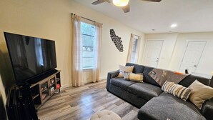 Un salon équipé d’un téléviseur à écran plat, d’un canapé, d’un pouf et d’un ventilateur de plafond.