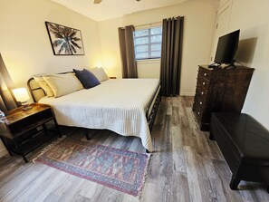 A bedroom with a bed, nightstand, dresser, TV, window with curtains, and a wall art piece. - Madrona's Cottage A- Vintage Arcade, Fenced Yard, Pets Welcome (North Charleston)