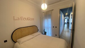 Een hotelkamer met een bed, een bord aan de muur met de tekst 'La Boutique', een plafondlamp en een gang die naar een andere kamer leidt.