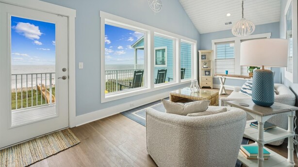 Amplia habitación con ventanas grandes que dan a una casa de playa, ventilador de techo y candelabro.