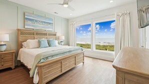 Un dormitorio con una ventana grande que ofrece vista a la playa, una cama de madera y un ventilador de techo.