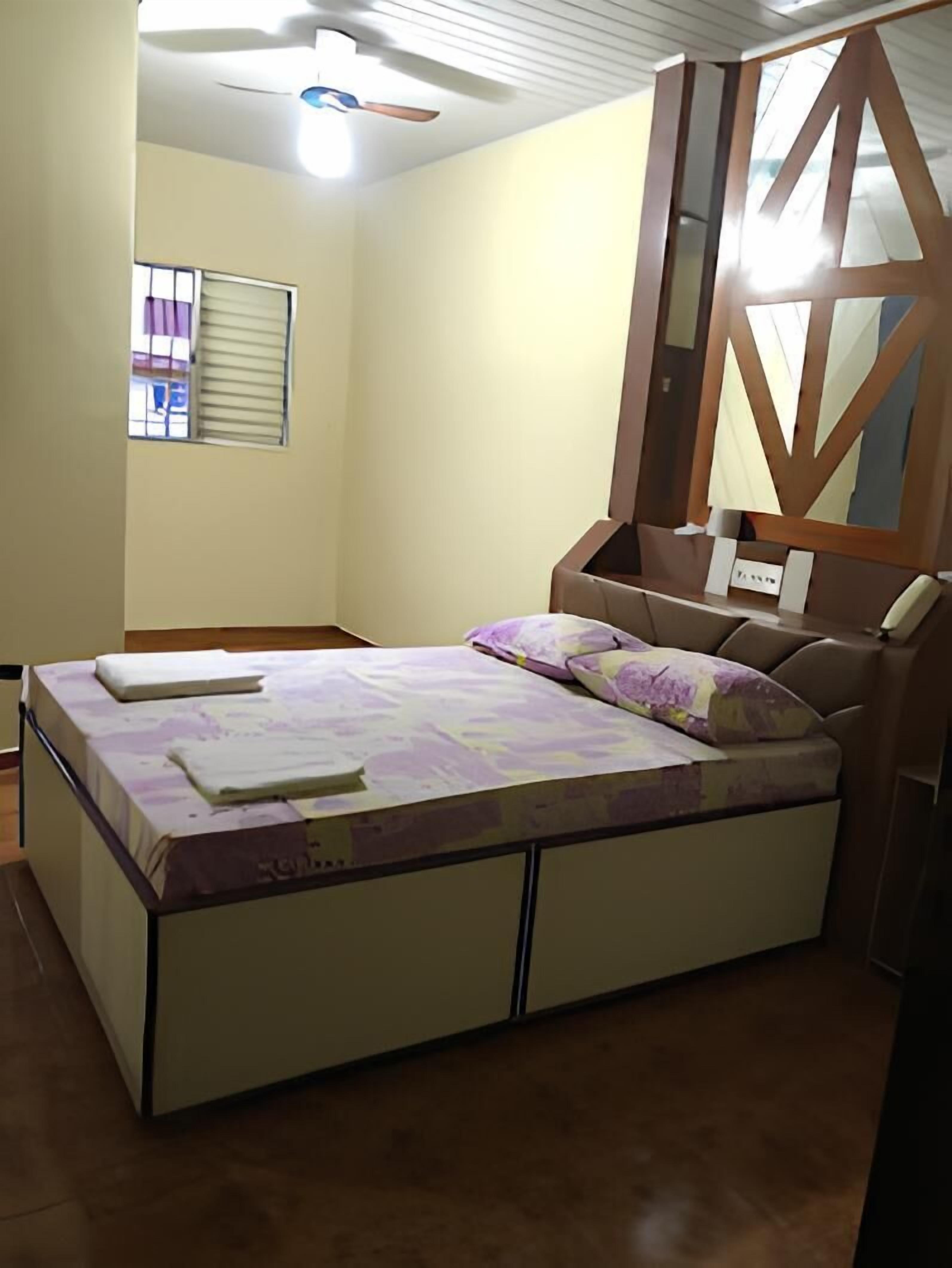 A bedroom with a bed, a ceiling fan, a window with blinds, and a nightstand with a lamp.