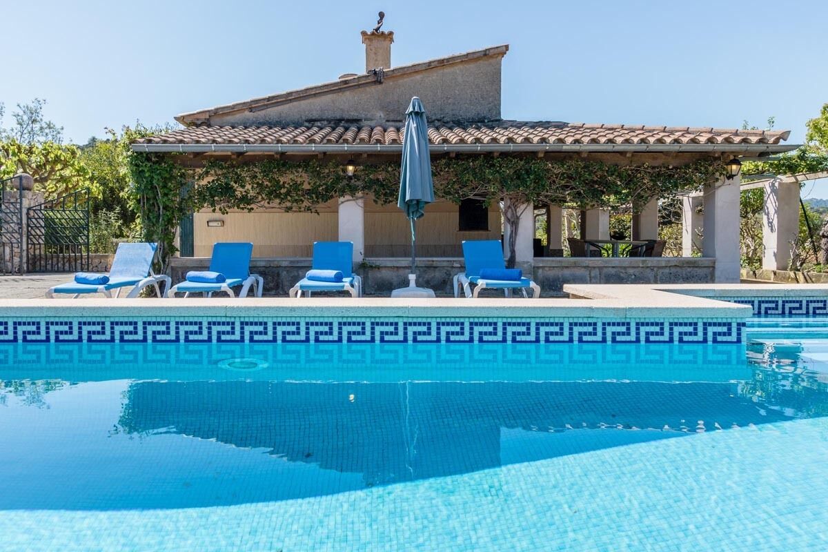 A pool with blue tiles and a decorative border, surrounded by lounge chairs and a pergola with a tiled roof.