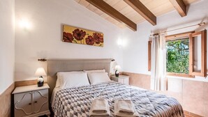 A bedroom with a checkered bedspread, a nightstand with a lamp, a painting of flowers on the wall, and a window with curtains. - H2924 Finca Catalina for 4 persons with pool near Pollenca (Pollenca)