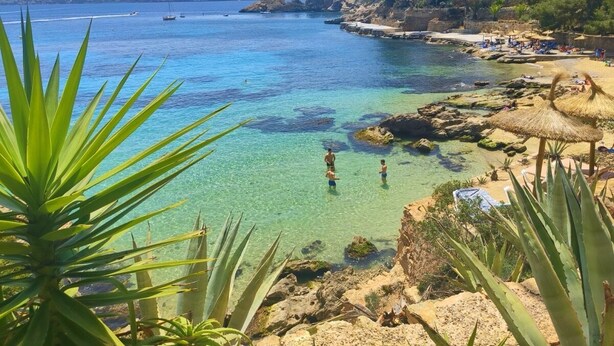 Vista costeira com águas cristalinas, litoral rochoso e uma praia com guarda-sóis.
