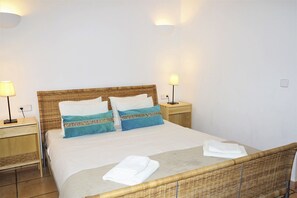 A neatly made bed with two pillows, a bedside table with a lamp, and a folded towel on the floor.