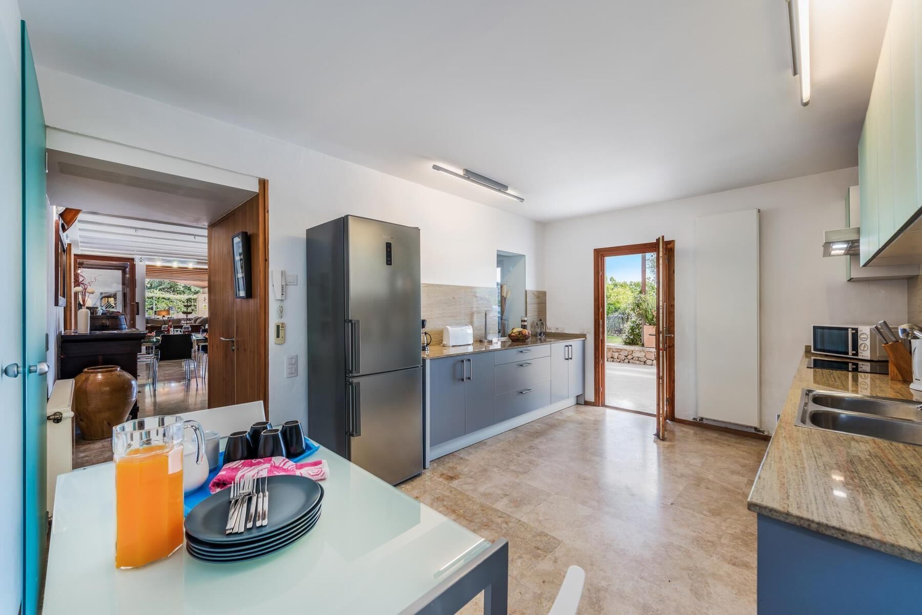 Een moderne keuken met een roestvrijstalen koelkast, een eethoek met een tafel gedekt voor vier personen, en uitzicht op de omgeving via een geopende deur.