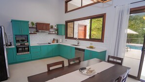 Eine moderne Küche mit türkisfarbenen Schränken, ein Essbereich mit einem Holztisch und ein großes Fenster mit Blick auf einen Pool.