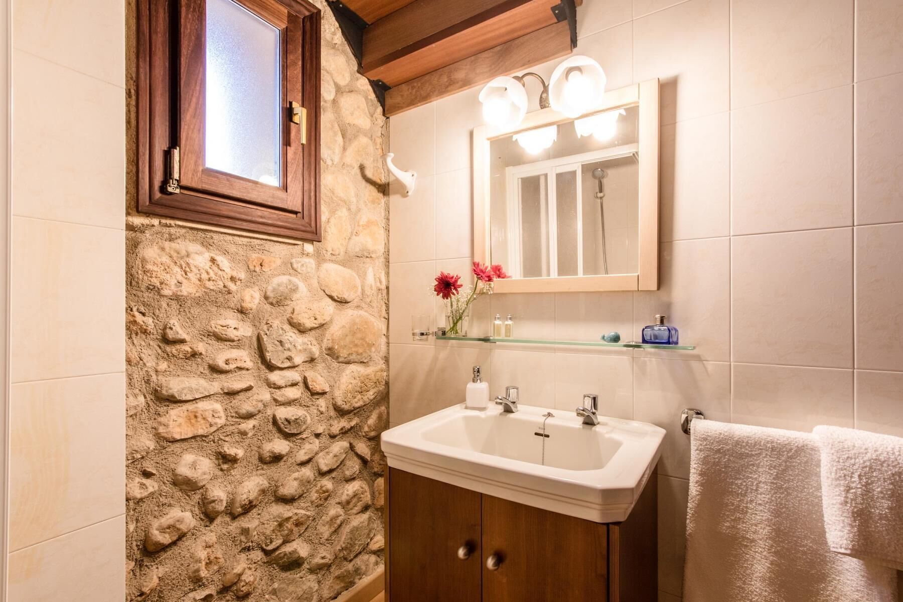 A bathroom with a stone wall, wooden cabinets, a white sink, a mirror with lights, and a towel rack.