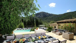 Uma área de refeições ao ar livre com piscina, mesas de jantar dispostas com pratos e copos, e vista para montanhas ao fundo.