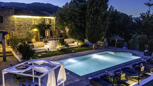 Uma área de piscina ao ar livre bem iluminada, com espreguiçadeiras, um espaço de estar coberto e um edifício de pedra ao fundo.